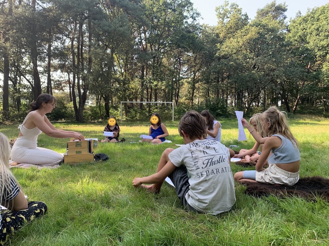 Foto van docent en leerlingen die op het gras zitten te zingen